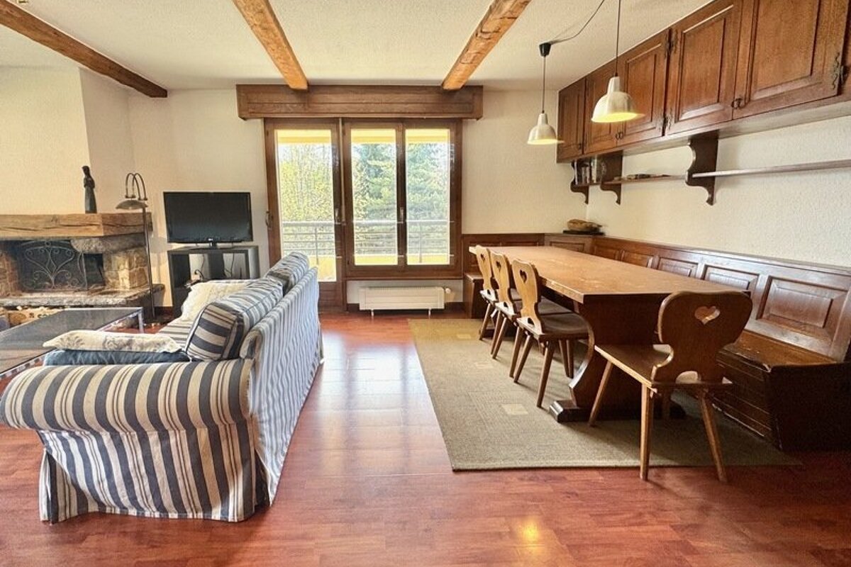 A rustic room featuring a striped sofa, a wooden dining table with bench seating, ceiling beams, and doors opening to a balcony.