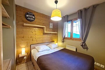 A cozy bedroom features a rustic wooden accent wall, a double bed, and a window with curtains. Warm lighting illuminates the inviting space.