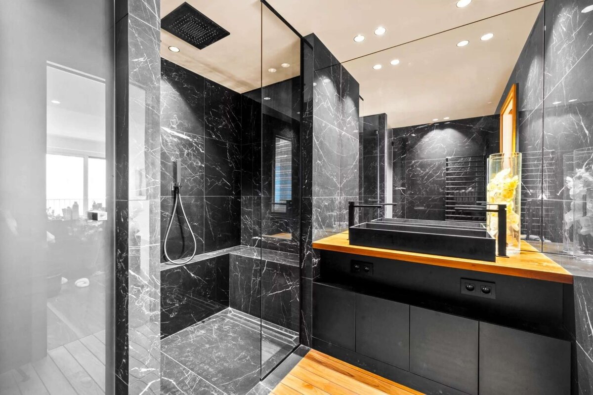Modern, luxurious bathroom showcasing striking black marble, a glass walk-in shower, a sleek black vanity with a wooden counter, and a matching wood floor.