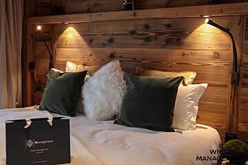 A cozy bedroom features a rustic wooden headboard, soft green and fuzzy white pillows, and warm reading lights, creating a comfortable ambiance.