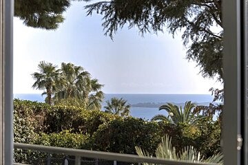 Scenic ocean view framed by a window, featuring lush green trees, palm trees, and a distant island under a clear sky.