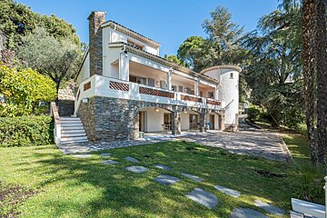 A multi-story white house with stone walls and a turret, surrounded by lush green trees and a sunny lawn with stepping stones.