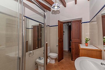 A traditional bathroom with a shower, toilet, bidet, and sink. It features wooden ceiling beams, terracotta floor tiles, tiled walls with blue accents, and rustic wooden doors.