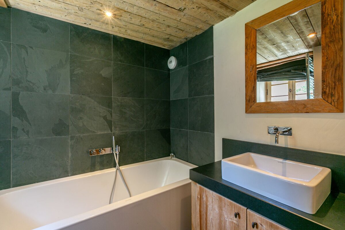 Rustic bathroom featuring a white bathtub with dark green tile walls and a wooden vanity with a vessel sink and mirror.