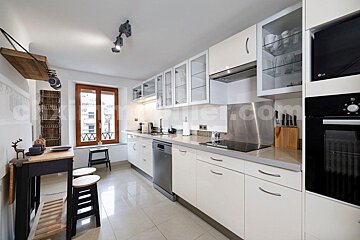 A modern white kitchen features sleek cabinets, stainless steel appliances, a window, and a compact breakfast bar with stools, lit by track lighting.