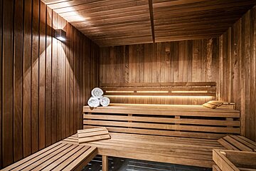 A warm, wooden sauna room with tiered benches, soft accent lighting, white rolled towels, and wooden headrests/mats.