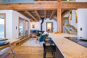 A cozy modern chalet interior featuring exposed wooden beams, a marble kitchen island with black stools, and an open-plan living space.