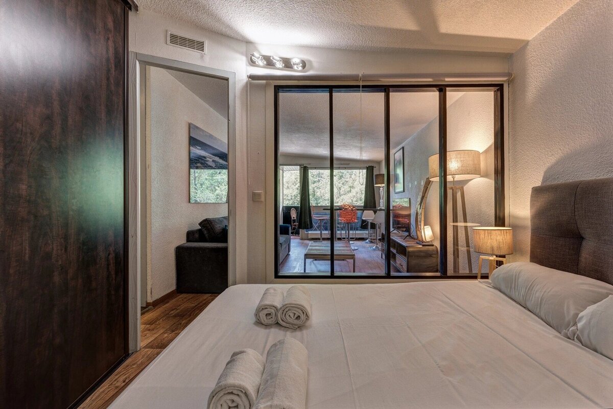 Cozy bedroom with white linens, rolled towels, and a glass wall looking into an adjacent living area.