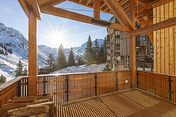 Sunny view of snowy mountains and pine trees from a rustic wooden chalet balcony.