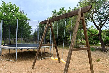 A wooden swing set with yellow swings and a trampoline are in a rustic backyard with trees and dirt ground.