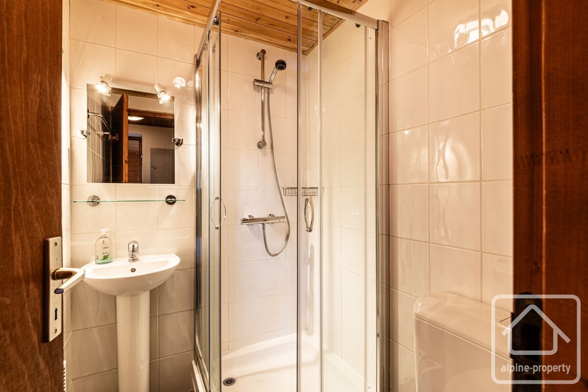 A small, functional bathroom with white tiled walls, a wooden ceiling, a pedestal sink, mirror, and a glass-enclosed corner shower.
