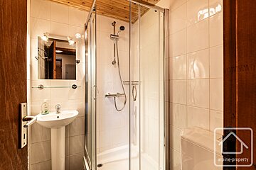 A small, functional bathroom with white tiled walls, a wooden ceiling, a pedestal sink, mirror, and a glass-enclosed corner shower.