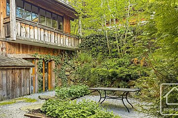 A rustic wooden house with a small gravel patio, featuring a metal table surrounded by lush green plants and trees.