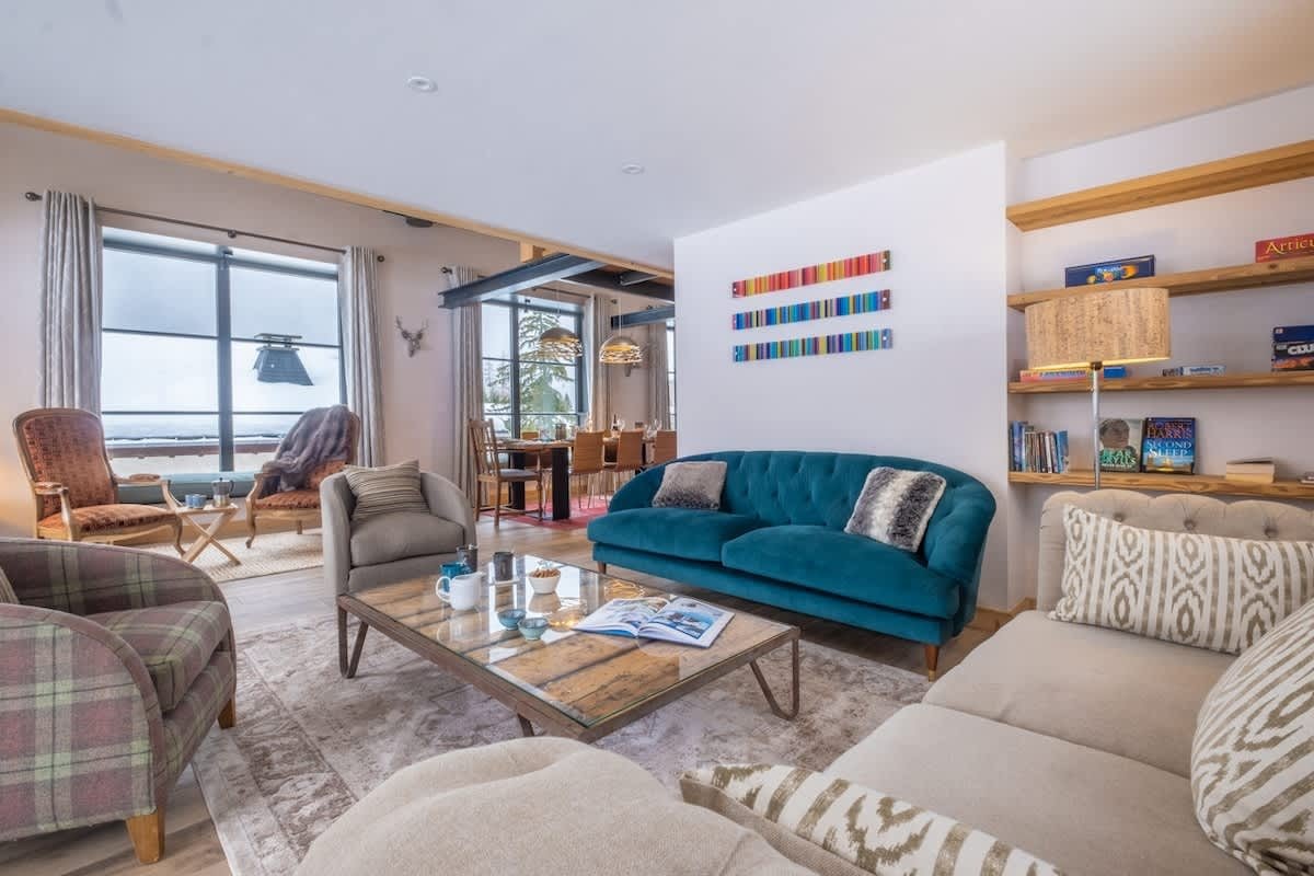 Cozy living room with a teal velvet sofa, patterned armchairs, glass coffee table, and large windows showing a snowy outdoor view.