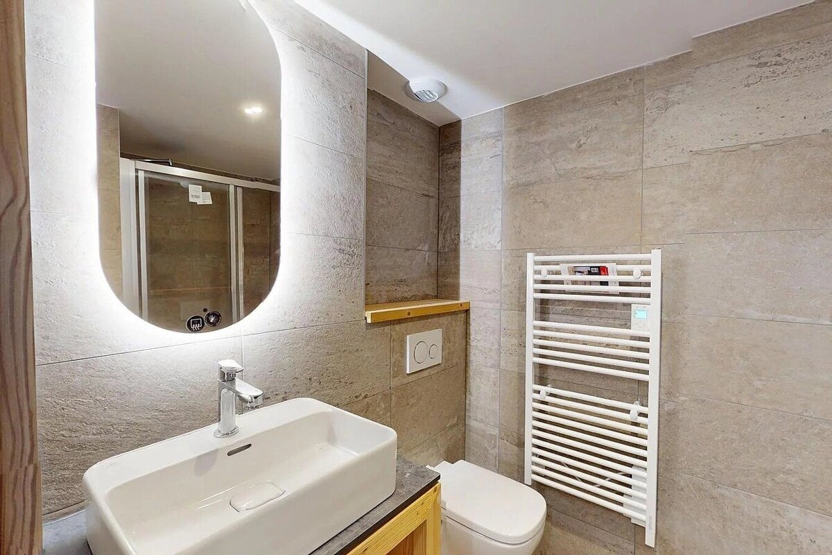 Modern bathroom featuring a backlit oval mirror, white vessel sink, toilet, beige tiled walls, and a white heated towel rail.