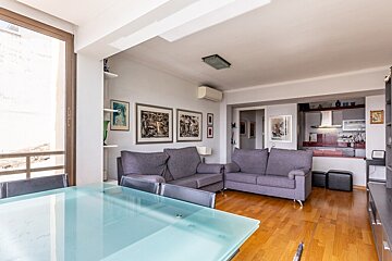Modern open-plan living and dining area with a glass table, purple sofas, hardwood floors, and a view into the kitchen.