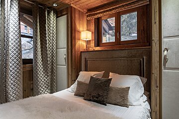 A cozy chalet bedroom with warm wooden walls, a plush bed, soft lighting, and a window showing an outdoor winter scene.