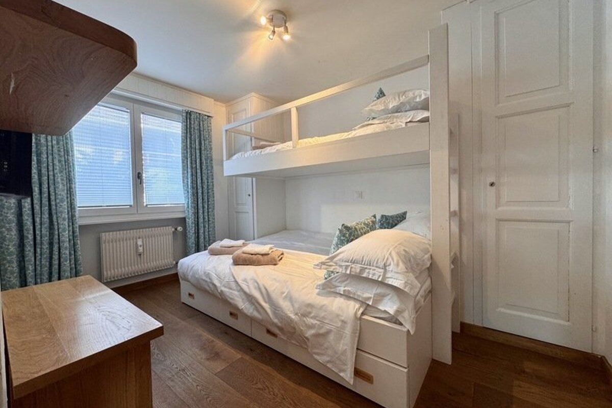 A cozy bedroom featuring a white wooden bunk bed, hardwood floors, built-in wardrobes, and a window with light blue curtains.