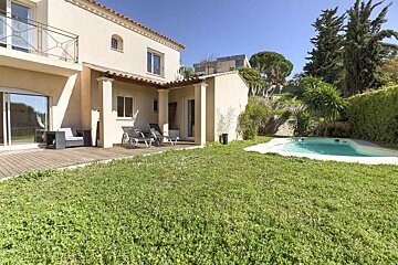 A beige villa with a patio, lounge chairs, a green lawn, and a small swimming pool. Hilly, treed background under a clear sky.