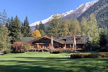 A large log cabin sits in the middle of a lush green field with mountains in the background