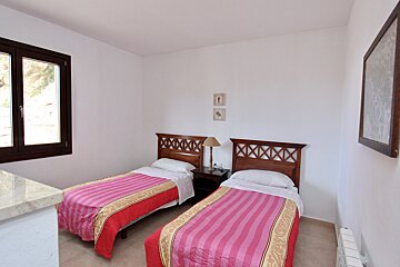 A bright bedroom with two single beds, wooden headboards, and pink/red striped blankets. A nightstand with a lamp, a window, and white walls are visible.