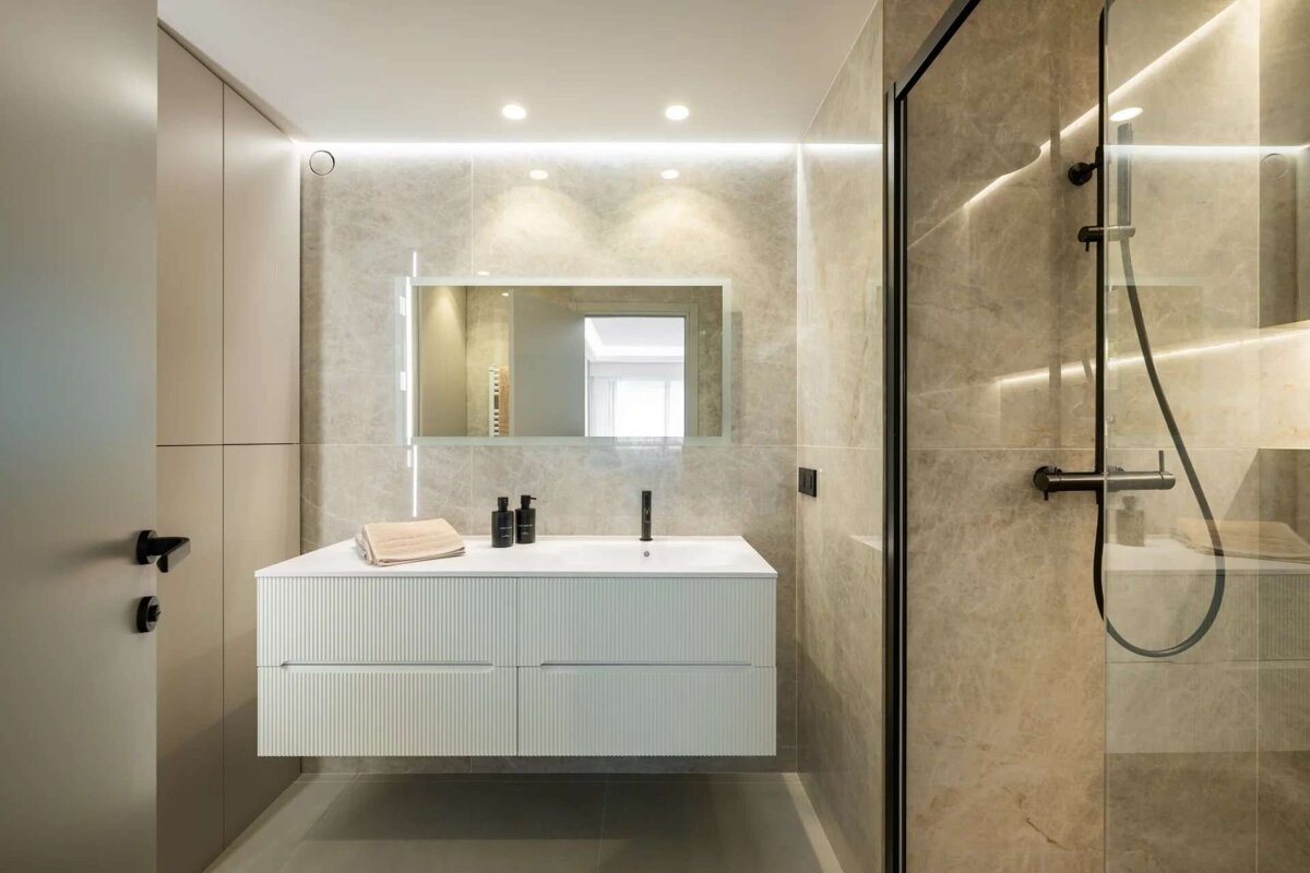 A sleek, modern bathroom with marble-look tiles, a floating white vanity, lighted mirror, and a glass shower. Subtle lighting creates an elegant ambiance.