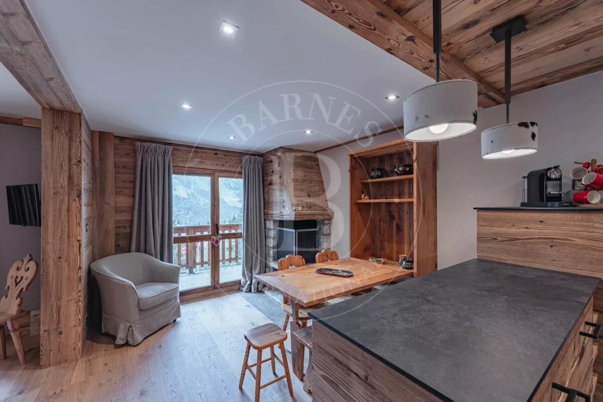 A wooden interior of a cabin with a dining table and kitchen island, looking towards glass doors with mountain views.