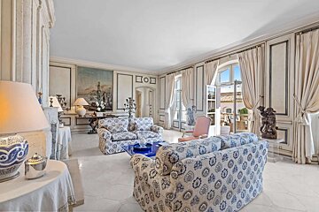 Luxurious white living room with blue patterned sofas, ornate details, and open French doors offering an outdoor glimpse. Bright and classic.