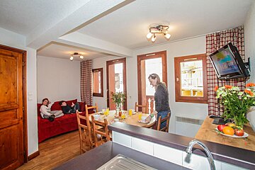A woman sets a dining table with juice and bowls, while two children relax on a red sofa in a cozy, rustic-style apartment living/dining area.