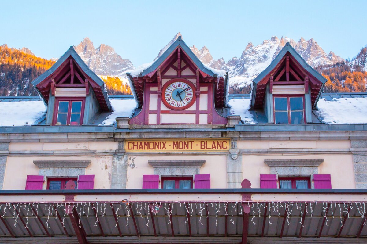 A Chamonix-Mont-Blanc building with a clock, snow-dusted roof, and festive lights, set against majestic, snow-capped mountains.