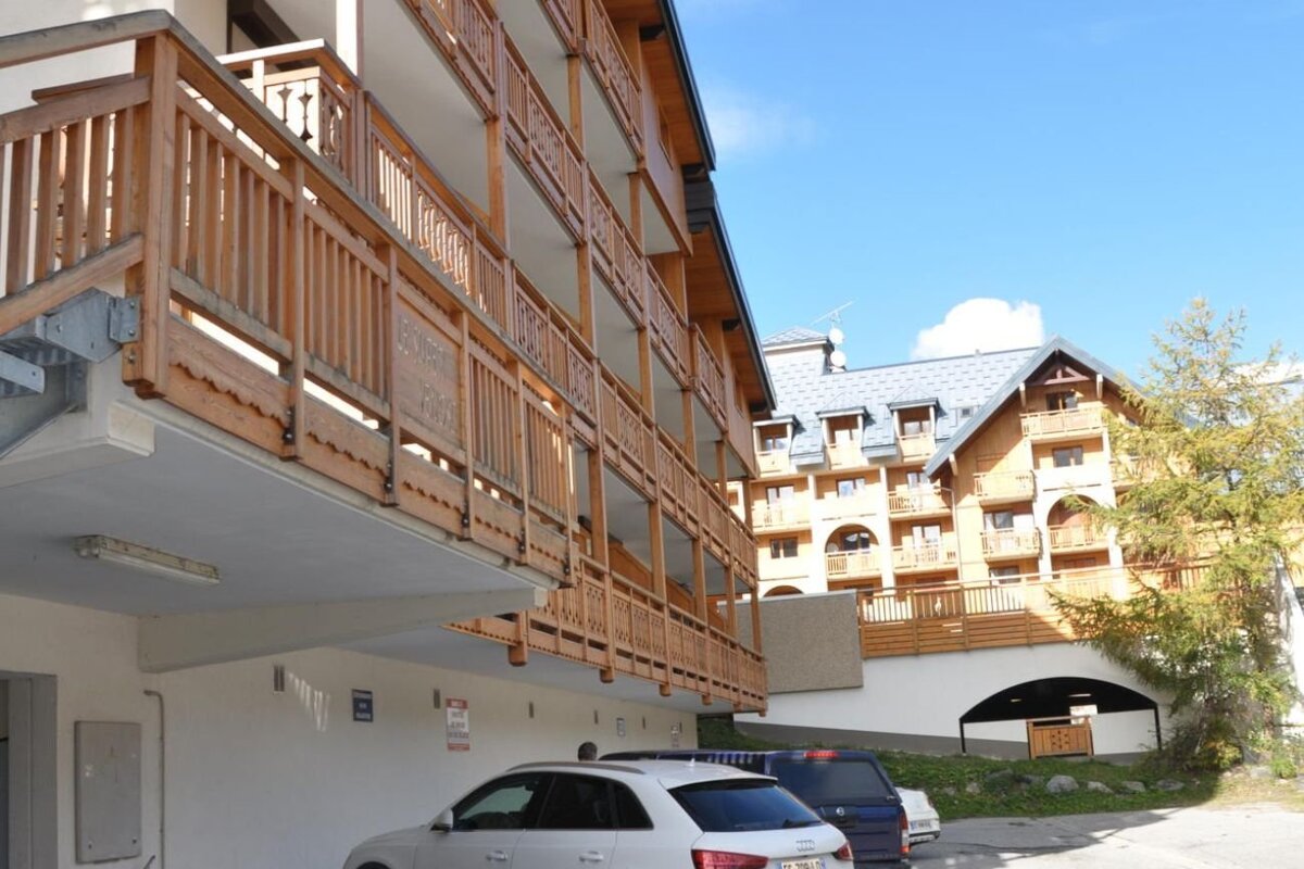 Multi-story chalet buildings with wooden balconies overlooking a parking area under a clear blue sky.