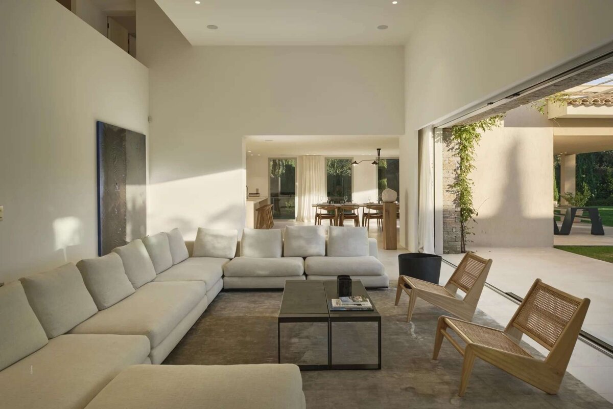 A chic, bright living room with a large cream sectional sofa, coffee table, and two wooden chairs. It connects to a dining area and an outdoor patio.
