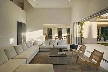 A chic, bright living room with a large cream sectional sofa, coffee table, and two wooden chairs. It connects to a dining area and an outdoor patio.