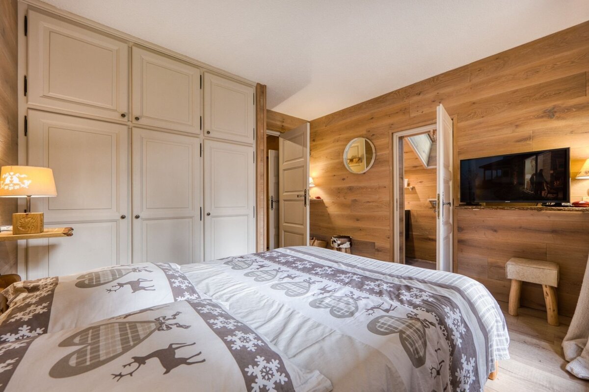 A rustic bedroom with light wood paneling, a large built-in wardrobe, a bed featuring reindeer and snowflake patterned bedding, and a mounted TV.