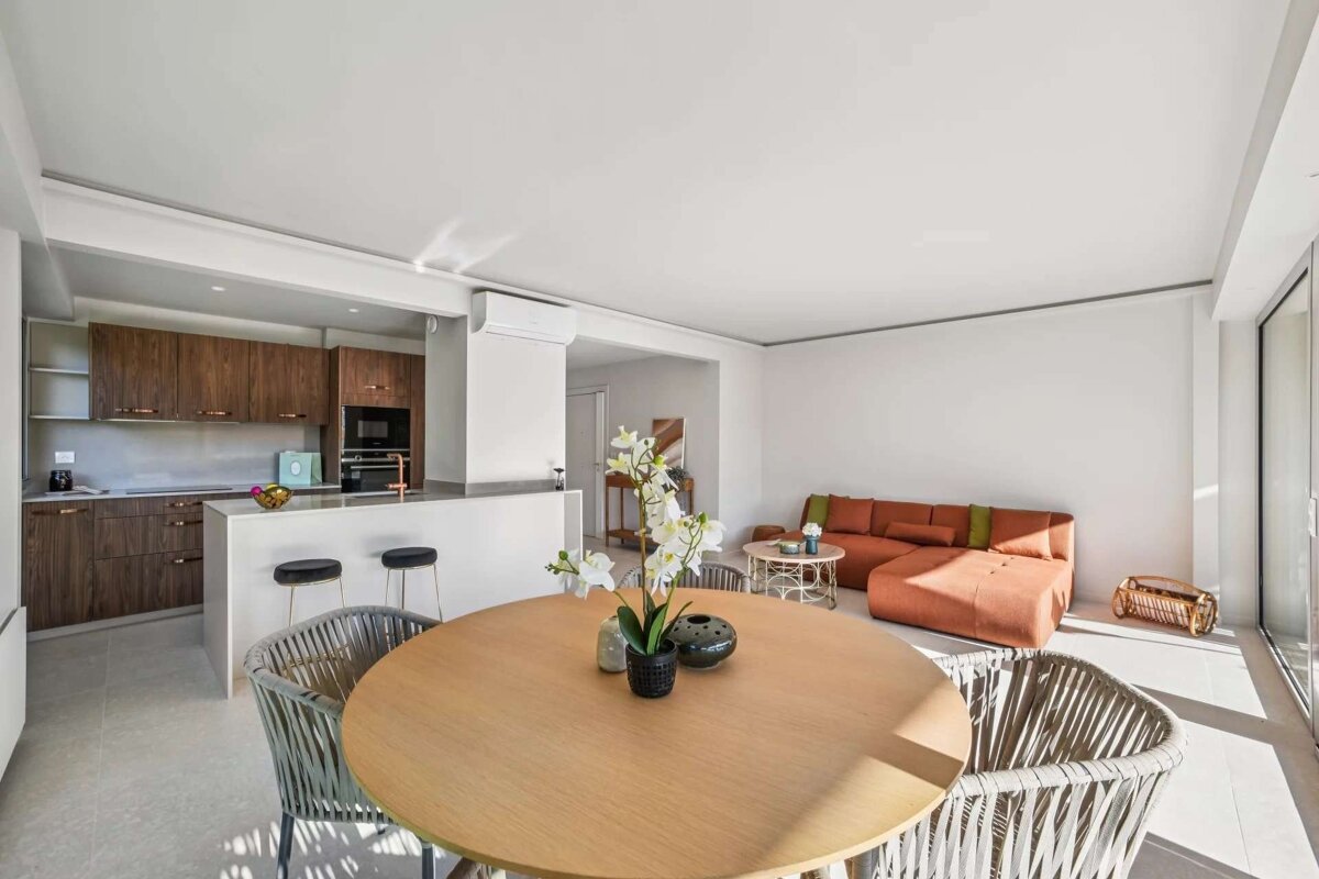 Bright, open-plan apartment featuring a modern wooden kitchen with an island, a round dining table, and an orange sectional sofa in the living area.