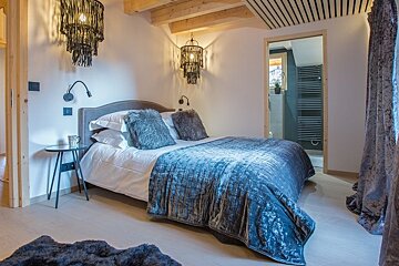 Cozy rustic-modern bedroom featuring a bed with a blue velvet duvet, fluffy pillows, wooden ceiling beams, and black fringe chandeliers.