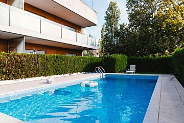 A clear blue swimming pool with an inflatable ring in front of a modern building, surrounded by lush green hedges and trees.
