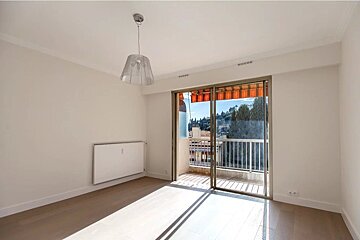 Bright, empty room with white walls, wooden floors, a hanging lamp, and sliding glass doors opening to a sunny balcony.