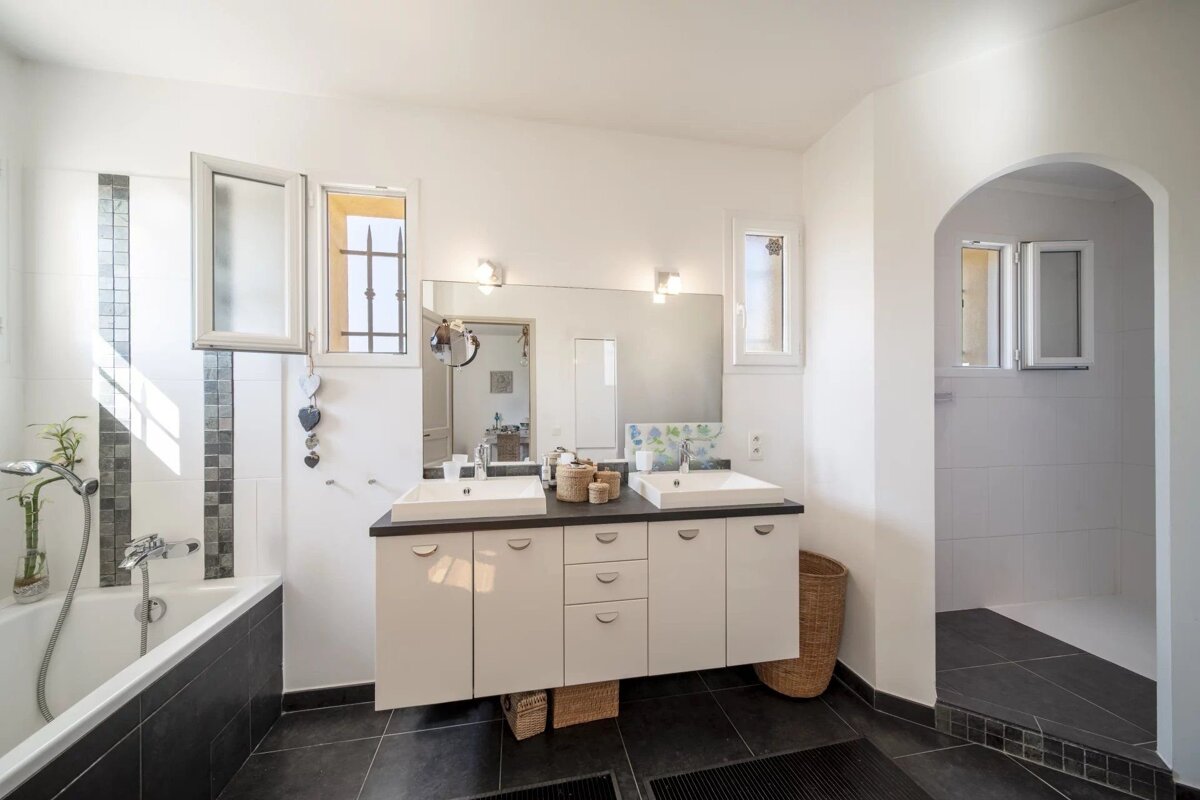 A modern white bathroom with dark tile floors, a double vanity, a bathtub, and an arched entryway to a shower.