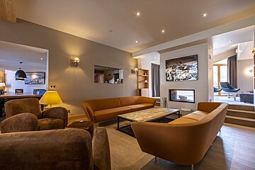A modern living room with warm earthy-toned sofas, a fireplace, wood floors, and soft lighting, leading to a dining area.
