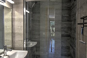 A modern bathroom with grey tiles, a glass shower cabin, sink, mirror, and a squeegee hanging on a rack.