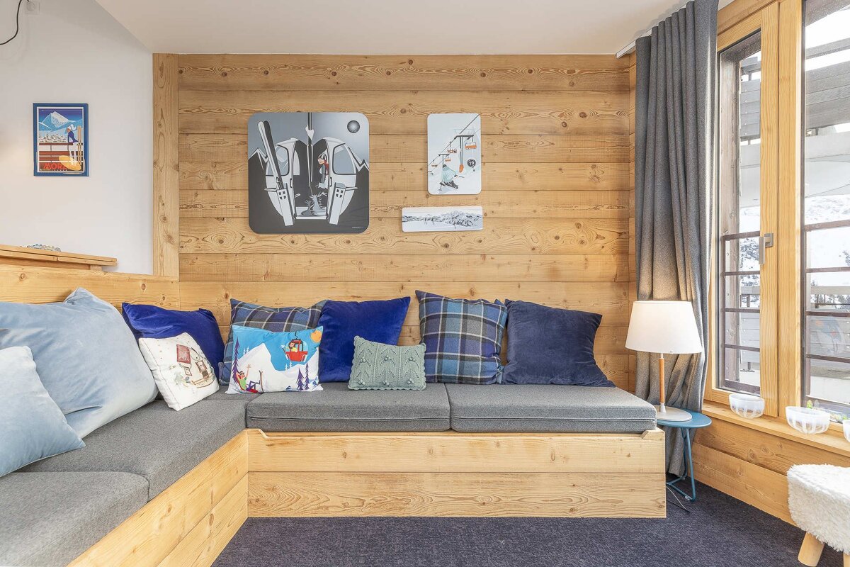 A cozy, L-shaped wooden sofa with blue cushions and ski-themed art on a wood-paneled wall, next to a large window.