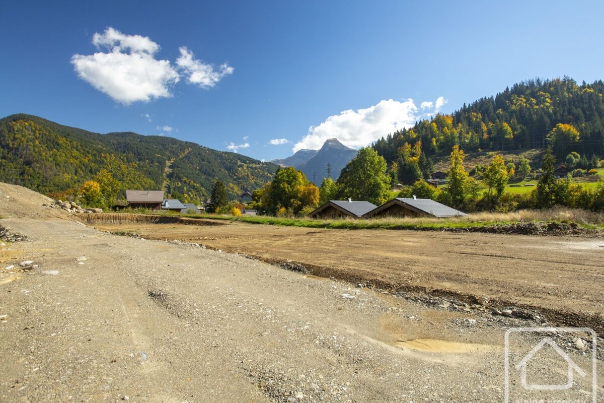 A gravel construction site in a sunny alpine valley, with houses nestled among autumn-colored trees and mountains under a clear blue sky.