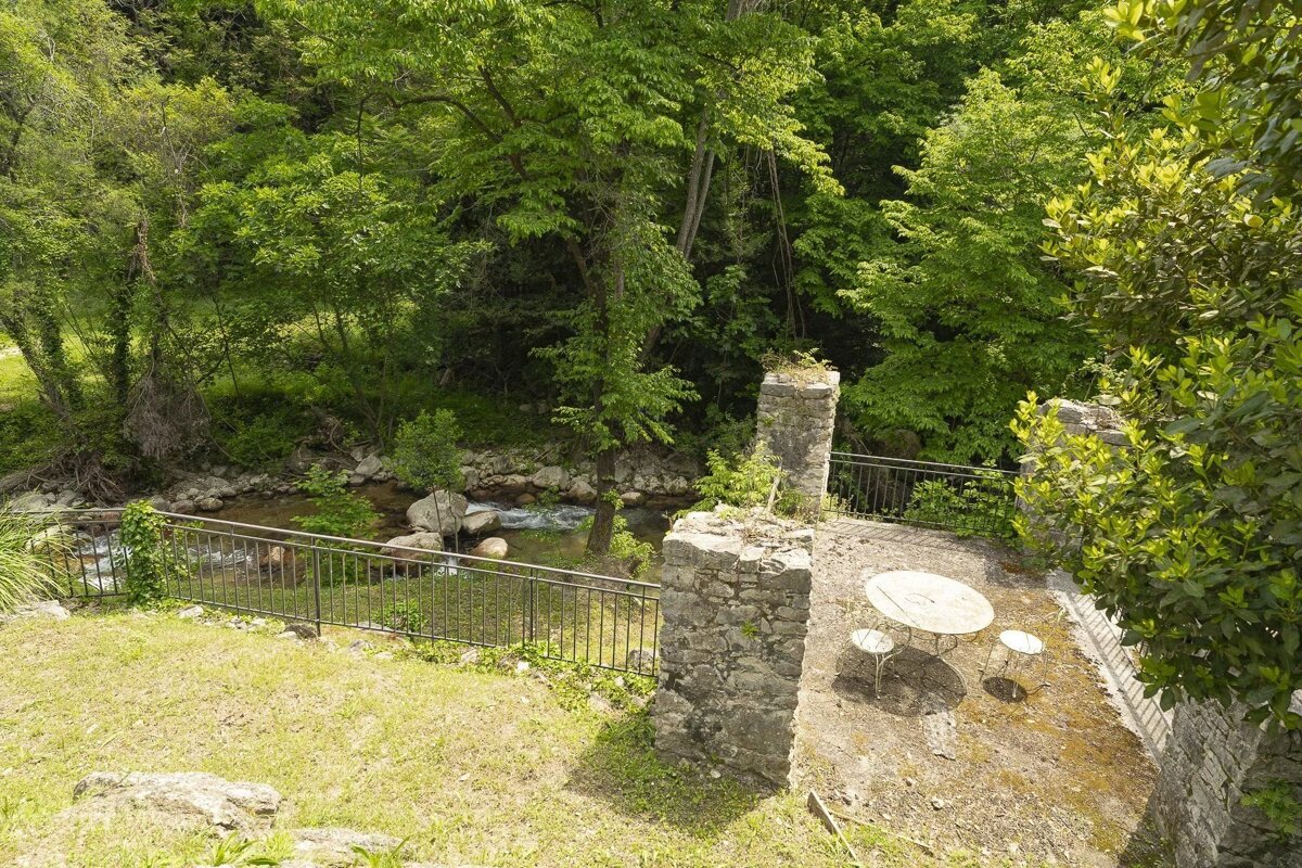 A stone patio with a table and chairs overlooks a lush green forest and a flowing stream, separated by a metal railing.