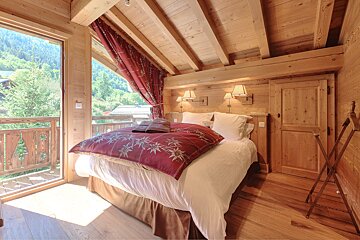 A rustic, all-wood bedroom features a large bed with a red floral duvet, red curtains, and a balcony overlooking a lush mountain landscape.