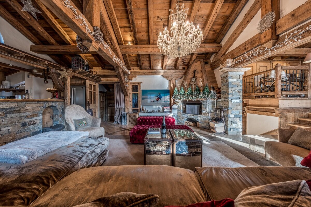 A lavish rustic ski chalet living room with exposed wooden beams, a grand chandelier, stone fireplace, and plush, inviting furniture.