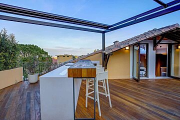 A modern rooftop terrace with a wooden deck, outdoor bar, and open room under a metal pergola, overlooking trees and a building with a tiled roof.