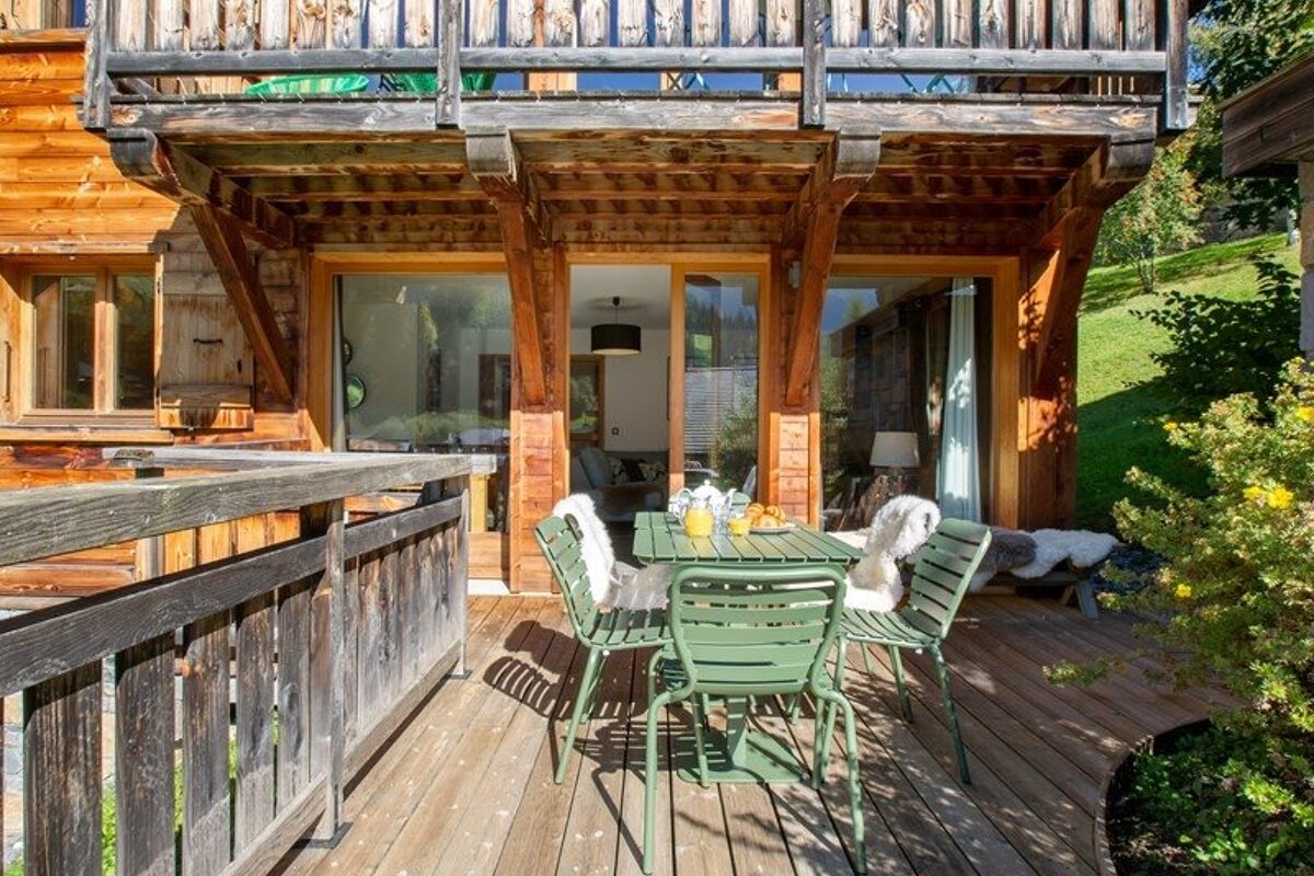 Wooden deck of a rustic chalet with a green metal dining table and chairs, set under an overhanging wooden balcony.