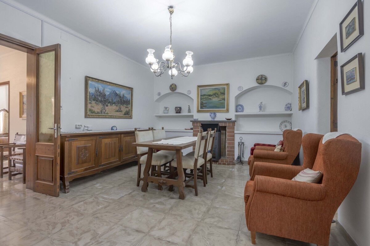 Rustic living/dining room with wooden table, orange armchairs, fireplace, and framed art on white walls. Chandelier and tiled floor complete the classic look.