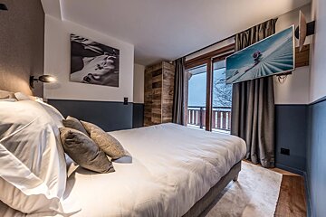 A chic bedroom with a comfy white bed, modern wall art, a TV showing a skier, and a balcony with a snowy mountain view.